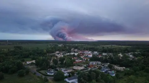 A done view showing the Sainte-Helene commune in France with smoke rising in the background during a wildfire in a forest area in Saumos