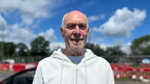 Julia Gregory / BBC A man, Lou Needle, wearing a white zip-up hoodie. He is stood in front of a road with roadworks next to it. 
