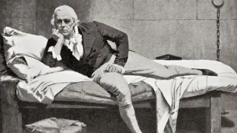 Getty Images A black-and-white painting of a man wearing early 19th Century clothes, including a buttoned, double-breasted dark jacket, a white ruffled shirt and light-coloured trousers, half-lying, half-sitting on a bed in a prison cell, his arm propping up his head. He has white hair and a reflective look on his face. The soft furnishings of the bed contrast with the stark surroundings, including stone walls and a metal chain hanging from the wall.
