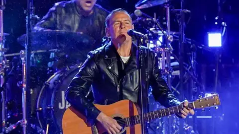 Bryan Adams performing onstage in 2022. He is holding an acoustic guitar and standing in front of a microphone. Behind him is a drummer and lots of blue lights. Bryan Adams is wearing a white top and black leather jacket. 