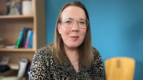 A woman with dark shoulder length hair, wearing glasses, a necklace and a leopard print shirt. There is a blue wall and a bookshelf behind her.