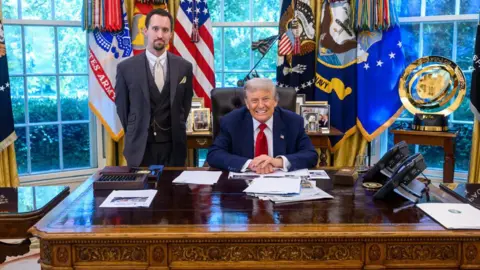 The White House is sitting behind a blue suit office and a red tie while Anotni stands in a gray suit of three pieces with a beige tie, white fitness and a series of watching in front of the windows covered with colored relationships and signs