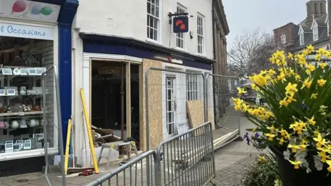 A view of the exterior of a Nationwide that has been damaged during a ram-raid incident. The front window has been removed and some cardboard blocks cover part of the gap. Metal fences surround the building. A bunch of yellow daffodils can be seen to the right of the image. 