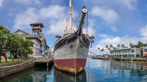 Getty Images A sailing ship, viewed from head on with mooring lines and dillapidated harbour buildings on the left hand side of the photo
