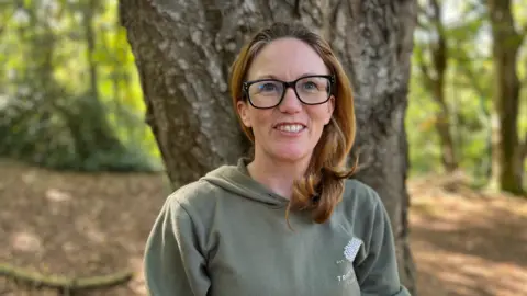 A woman in a green hoodie wearing glasses and standing in the woodland.