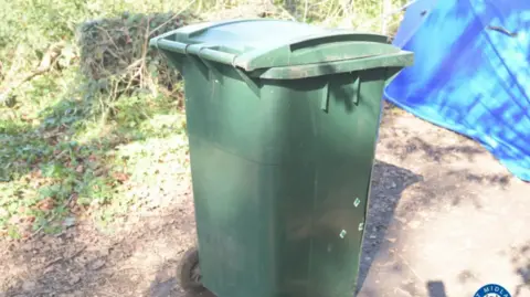 West Midlands Police A green wheelie bin on a path with bushes and a blue tent in the background