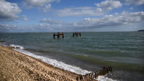 A sunny beach scene with blue skies and a few white clouds. Two old looking rusty structures are popping up from the water in the middle of the image. The beach is stony.