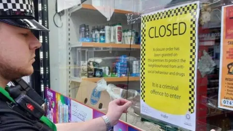 Cambs Police Police officer pins a notice to a shop window which says it is an offence to enter the shop which has been issued a closure notice. The notice said this was being carried out to protect the community from anti-social behaviour and drugs