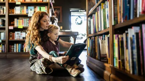 A stock image of a woman with curly hair reading a story to a young boy sitting in her lap. They are in a library. 
