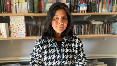 Melissa Esquerre wearing a white and black checked fleece with a blue zip. She has dark, shoulder length hair and is staring straight at the camera. She is standing in front of two shelves filled with books and DVDs. The spines are a multitude of colours.