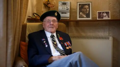 A very old man in a decorative army uniform. He sits in a chair wearing a blazer and beret. The blazer is adorned with war medals and a poppy. A radiator and shelf with photos and a small hi-fi system can be seen behind him.