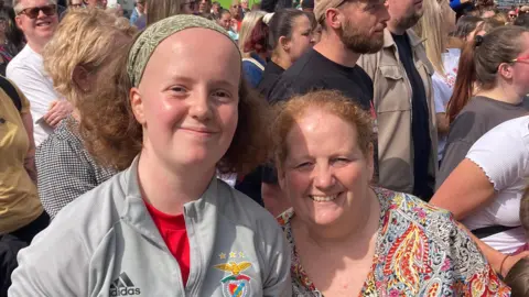 Justin Dealey/BBC Sandra and Lana are both smiling amongst the crowd. Lana is wearing a green headband and a grey sports top. Sandra is wearing a colourful V-neck top.