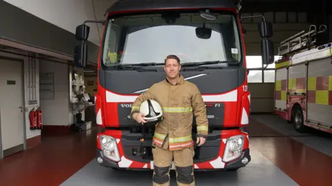 Jonner Robinson is standing in front of a fire engine in his uniform with his helmet tucked under his arm. He is 38 years old with short dark hair 