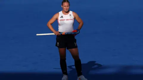 Getty Images Hockey player Sarah Robertson in Great Britain white top and navy skirt stands with hands on hips, and holding hockey stick, with blue pitch in background.