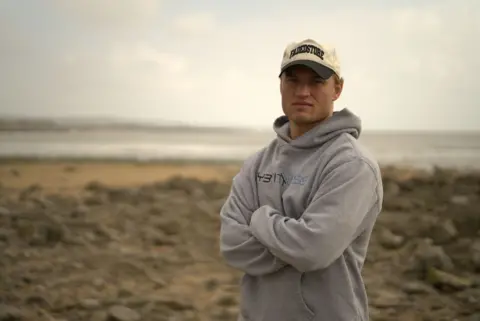 BBC Hari Morgan standing with his arms crossed, wearing a cap and grey hoodie on the beach.