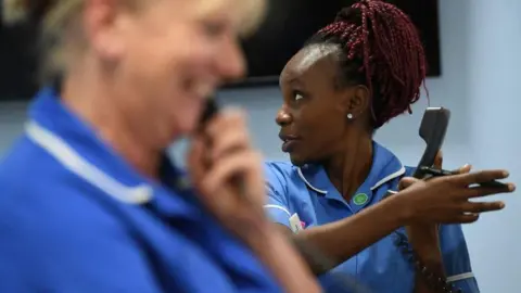 Two female nurses in blue uniforms holding landline phones. One is listening to the person on the other end of the phone, the other is holding the phone away from herself and talking to someone else.