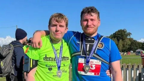 Two men stand, arm in arm, next to each other smiling at the camera. They both have medals round their necks. The man on the left is wearing a neon green shirt with Shrewsbury half marathon written on it. The man on the right has a blue shirt on with a red race sticker and black phone holder on. There is a blue sky in the background and a green field with fences. 