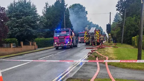 Pete Benwell Several fire engines on a road, with fire hoses running along the verge.
