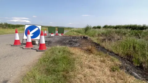 A burnt patch of road surface and grass at the side of a minor road in the countryside. The sky above is blue. There are cones and road signs surrounding the burnt patch.