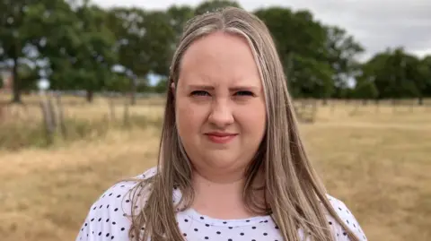 Simon Spark/BBC Holly McLeod. She has long fair hair and is wearing a spotted top. She is standing in a field with trees in the background.