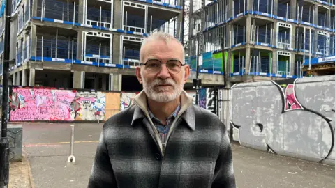 Andrew Boff, chair of the planning and regeneration committee for the London Assembly outside a construction. The block is still built so you can see the exposed framework, and at ground level are two hoardings he's standing in front or, with graffiti on them.