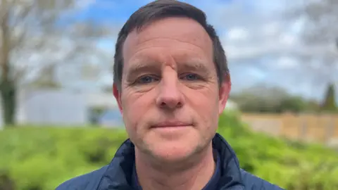 A man with short dark hair, looking into the camera. He is standing outside, a green bush, trees and blue sky can be seen in the background, albeit out of focus. The man is wearing a blue coat. 
