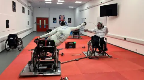 Dimitri Coutya on the right, sparring with his opponent on the left. They are in the training facility room at Bath University. It is a long rectangular room with red padding on the floor. Both fencers are sitting in their wheelchairs wearing their white protective kit and fencing hoods with the GB flag on them. They are leaning out of their wheelchairs to try and strike each other with their fencing foils (swords) 