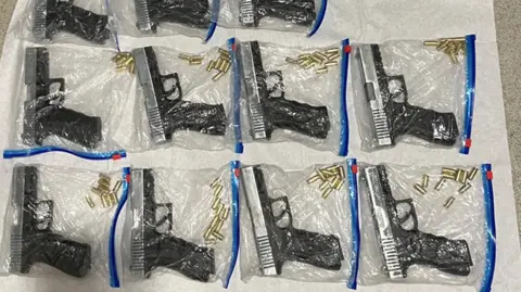 ERSOU 11 small black guns all contained inside zip lock bags with gold bullets. They are laid out on a white sheet on the floor.