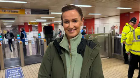 BBC/Jim Scott Sgt Louise Page, who is wearing a green fleece with a gold zipper and a large green overcoat, is standing in front of the ticket gates at Gateshead Metro station. To the right of her are two police officers, who are wearing yellow high visibility coats, which say 'POLICE' in blue. 