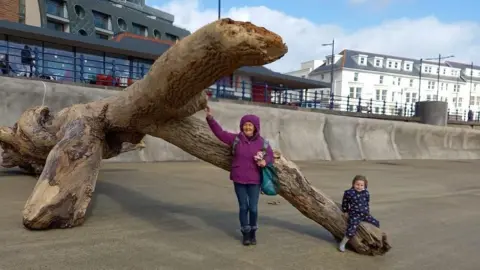 Chris Kitney Photograph showing the large piece of driftwood, named Lizzy. Izzy, aged six, sits on the forefront of the piece of wood, while her grandmother stands near the centre. 