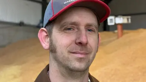 Farmer Philip Weston stands in a barn full of grain. he has sideburns and wears a blue baseball cap.