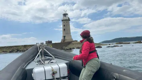 Evie Smith Jennifer Kloester on a small black boat on water looking out towards the lighthouse. She is wearing a red jacket, red cap and green trousers. The engine of the small boat is grey and white. To the right, at the end of the breakwater, is the lighthouse which is grey.