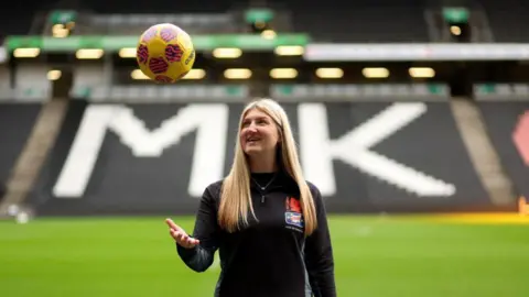 A woman with long straight blonde hair wearing a dark tracksuit throwing a yellow football in the air with her right hand. She is standing in MK Dons' stadium and the letters "MK" can be seen in a stand behind her. 