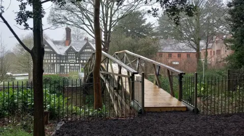 National Trust/David Bennett The restored bridge with railings on either side of it and buildings in the background. The wooden bridge is arched and has a new shiny base. 