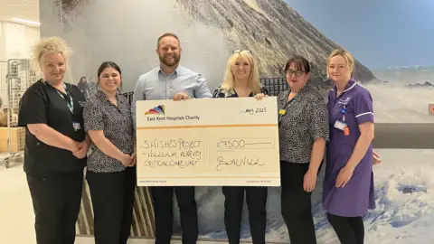 A group of people stood holding up a large cheque in a hospital corridor. 