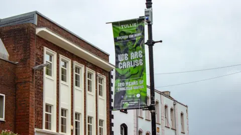 Tullie A green banner attached to a black lamppost with a photo of men hugging. The banner reads "Tullie- Backing the Blues- We Are Carlise- 120 years of Carlisle United." Carlisle is spelled with one l in the words 'We are Carlise'.