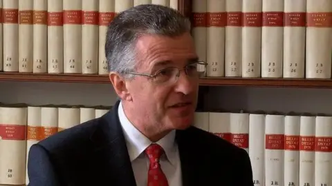 Richard McMahon, who has grey hair and wears glasses, looks off to the side with a row of white and red books behind him. 