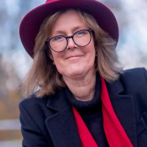 Ann Russell A woman wearing glasses, a dark blue blazer, a red brimmed hat and a red scarf smiles at the camera.