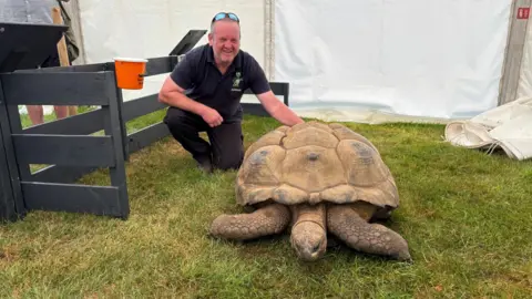 Luke Deal/BBC A smiling man kneels on one knee beside a giant tortoise
