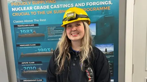 A women in her early 20s with long blond hair is smiling. She is wearing a hard hat and has a pair of safety glasses in the top pocket of her black jacket. She is stood indoors in front of a display with facts and figures about Forgemasters