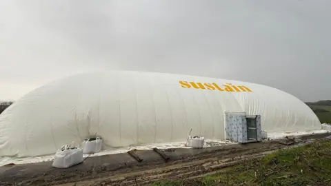 A large white inflated dome made from tent-like material. It is rounded, tall and in an oblong shape with an entryway. The word Sustain is printed on it in yellow. It sits on a muddy field under a grey sky.