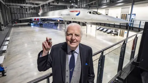 PA/Ben Birchall John Britton, Concorde's chief engineer, holds the new coin in his uplifted hand, standing on a platform in the hangar with Concorde behind him. He is smartly dressed in a blue jacket, striped shirt and tie. 