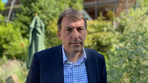 A middle-aged man stares at the camera wearing a shirt and jacket with a sun umbrella and trees in the background.