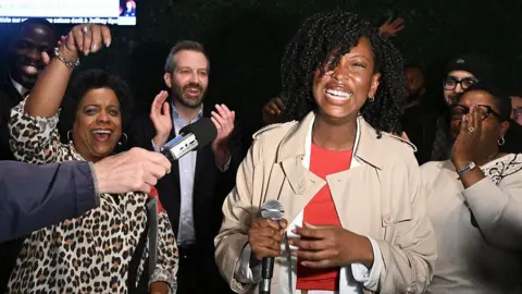 Bloomberg via Getty Images Tatiana Auguste, wearing a beige trenchcoat and a bright red sweater, smiles widely as she holds a microphone. She is surrounded by a crowd of celebrating supporters. 