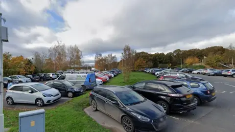 A large car park that is full of cars. There is a small strip of grassland in between two sections of the car park with trees in the distance. 