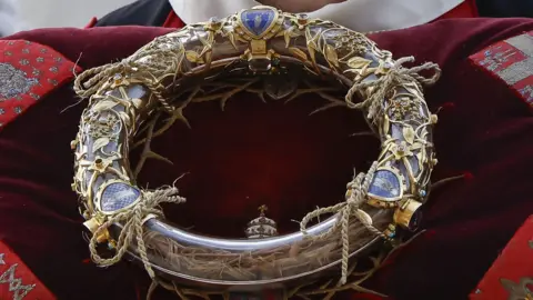 A close-up of the Crown of Thorns encased in a transparent tube with gold decorative details, placed on a red cushion. 