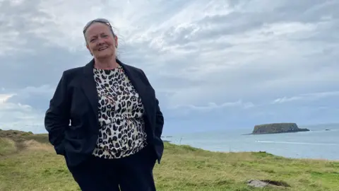 Suzanne Buckner stands on a grassy clifftop with the sea and a small island in the background. She wears a black-and-white blouse, dark trousers and a dark jacket, with sunglasses pushed back over her brown hair.