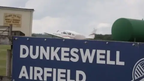 A plane taking off at Dunkeswell Airfield. There is a white plane with a blue sign in front of it.