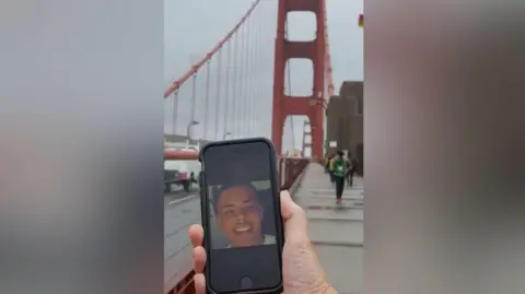 Family Breon pictured at The Golden Gate Bridge in California. We see a hand holding an iPhone with a picture of him. The landmark is in the background. It is a gloomy day and there are people walking up and down but they are blurry.