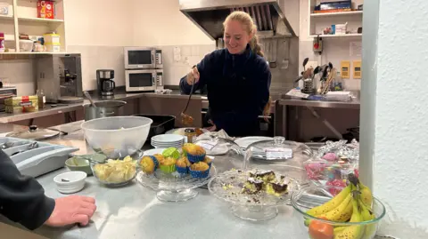 King's Arm Project A woman, in a kitchen, putting food on a plate, with a ladle in her right hand, standing in front of a kitchen with a cooker, microwave and utensils. A large amount of food is in front of her, including cakes, bananas and bowls. 